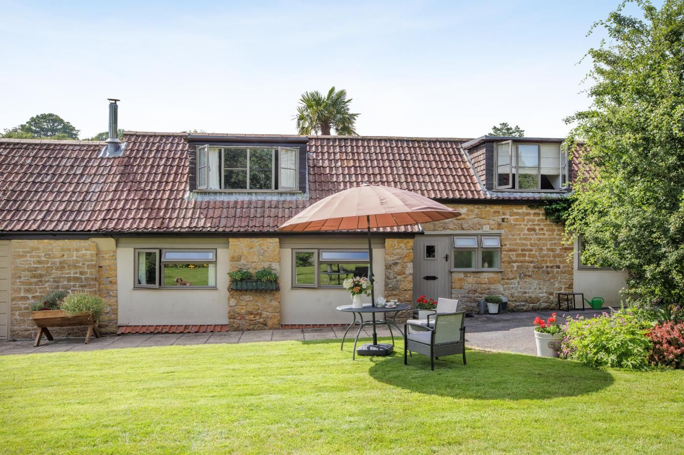 A house with a tiled roof, stone walls, a lawn, a patio with a table and chairs, and a window with a view of the garden.
