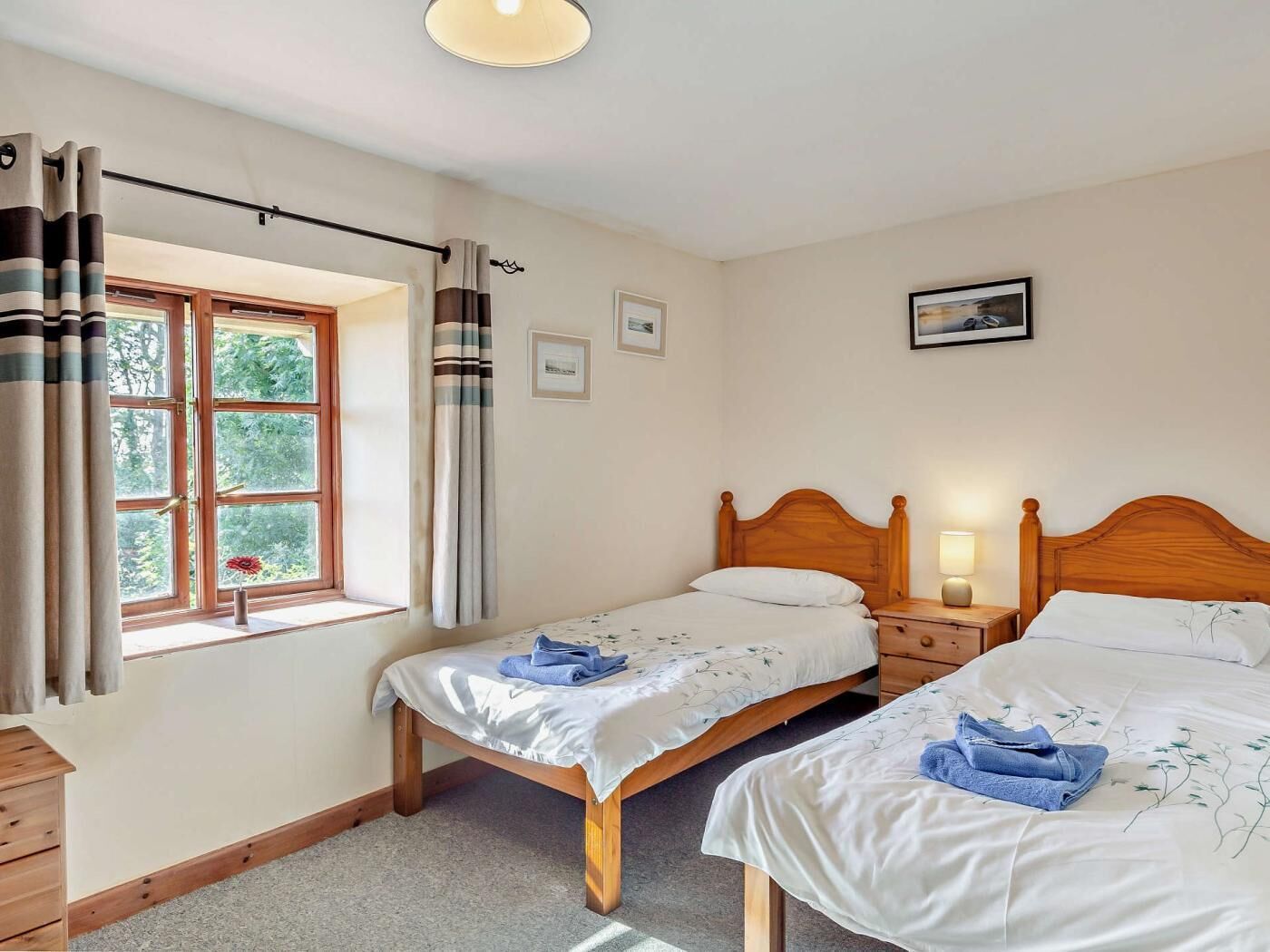 Un dormitorio con dos camas individuales, una ventana con cortinas, una mesita de noche con una lámpara y un cuadro enmarcado en la pared.