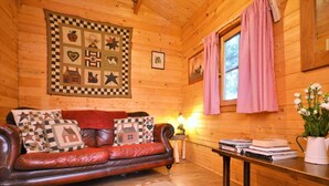A cozy log cabin interior with a leather sofa, quilted wall hanging, and a window with curtains.