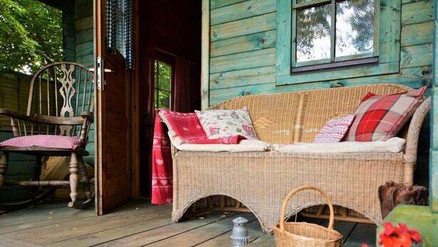 A rustic wooden porch with a wicker sofa, a rocking chair, a lantern, and a basket.