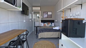 Quarto de hotel compacto com cozinha compacta, cama com cobertura cinzenta, televisão e pequena área de refeições.