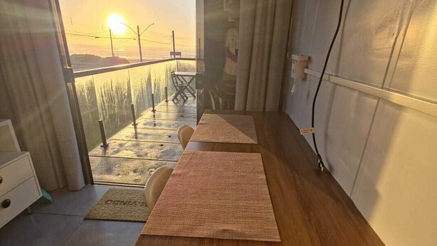 Quarto com escrivaninha de madeira, uma cadeira, tapete tecido e vista do pôr do sol por uma janela grande.