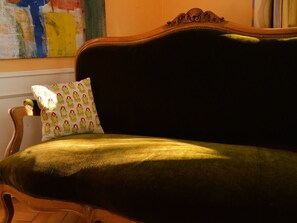 Een groene gestoffeerde bank met een gekleurd kussen, in een kamer met een houten stoel en een schilderij aan de muur.