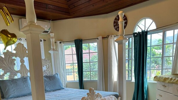 Une chambre à coucher avec un lit à baldaquin, une commode blanche et une vue sur l’extérieur grâce à de grandes fenêtres.