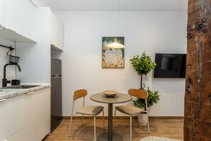 A modern kitchen with white cabinets, a stainless steel refrigerator, a dining table with two chairs, a hanging pendant light, and a flat-screen TV on the wall. - A studio in the heart of Malasaña's creative quarter, where every corner has been composed like a gallery you get to live in. (MADRID)