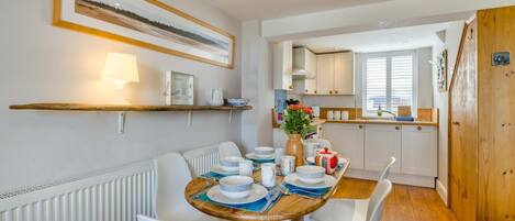 Un comedor con una mesa redonda dispuesta para cuatro comensales, sillas blancas, piso de madera, radiador blanco, un estante con una lámpara y un cuadro en la pared.