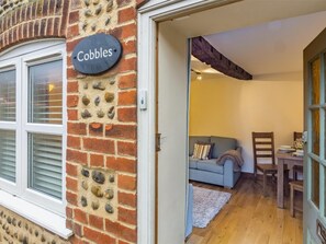 Ein Ziegelhaus mit einem 'Cobbles'-Schild, einem Fenster mit Rollläden und einem Blick in den Innenraum mit Sofa und Esstisch.