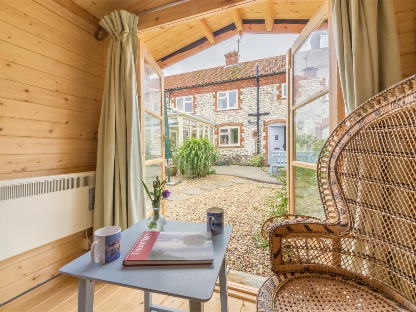 A wooden interior with a table, a wicker chair, and a view of a stone building outside.