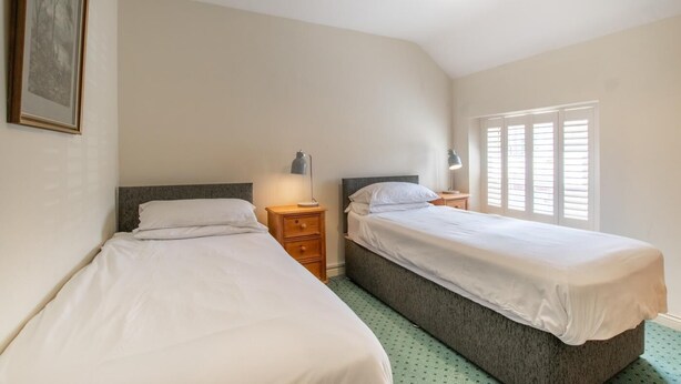 Un dormitorio con dos camas individuales, una mesita de noche con lámpara, un cuadro en la pared y una ventana con persianas.