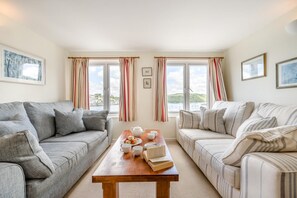 Ein Wohnzimmer mit zwei Sofas, einem Couchtisch mit Buch und Teekanne, und Blick auf das Meer durch Fenster.