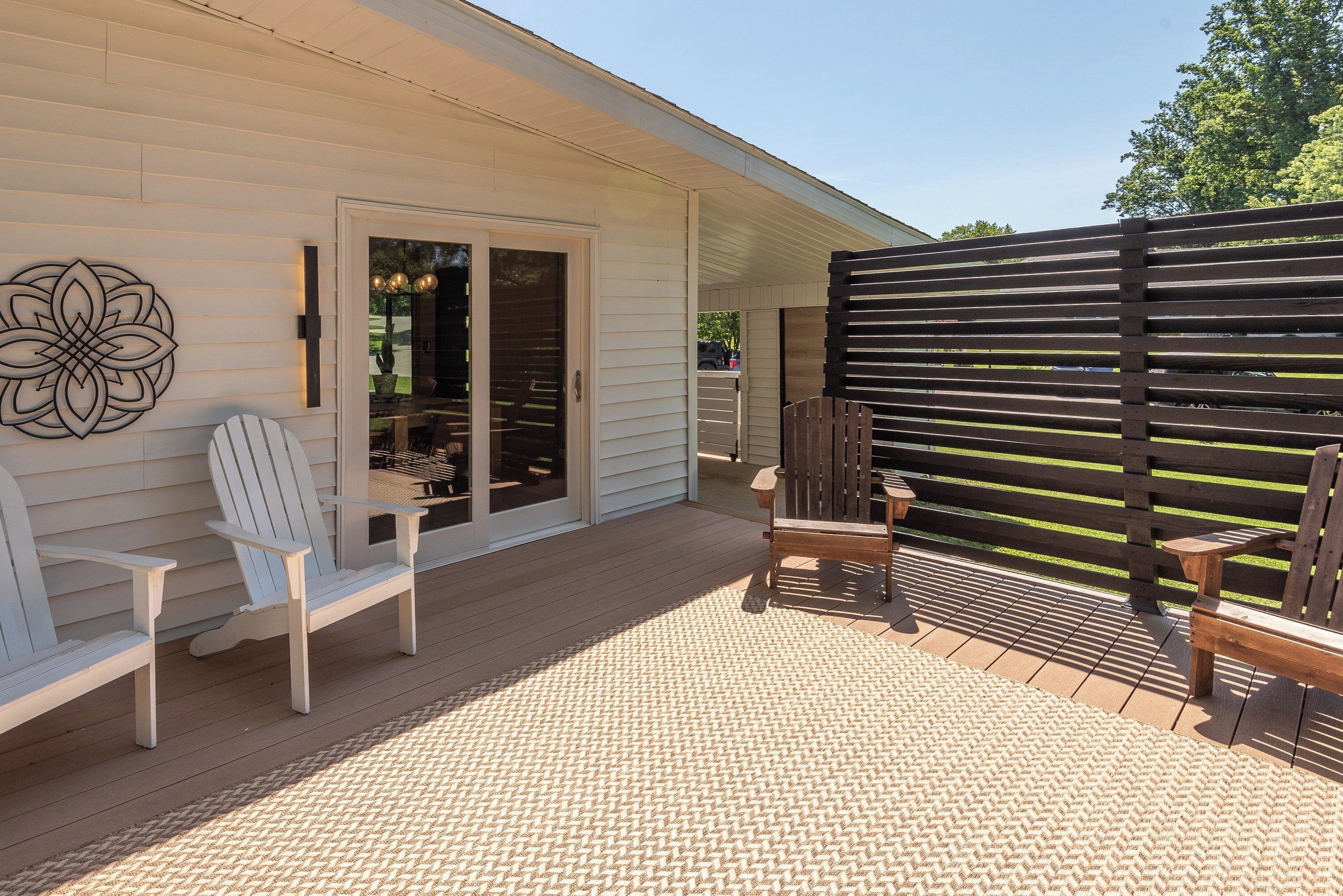 A covered patio with a wooden deck, two Adirondack chairs, a woven rug, and a decorative wall piece.