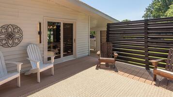 A covered patio with a wooden deck, two Adirondack chairs, a woven rug, and a decorative wall piece.