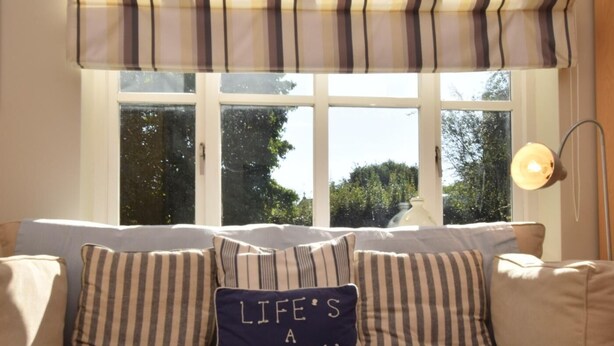 Ein Sofa mit gestreiften Kissen und einem Kissen mit der Aufschrift 'Life's a Beach', ein Fenster mit Blick auf Bäume und eine Lampe rechts.
