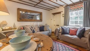 Een gezellige woonkamer met een houten plafond, een bank, een eettafel met een schaal en bestek, een spiegel en een raam met gordijnen.
