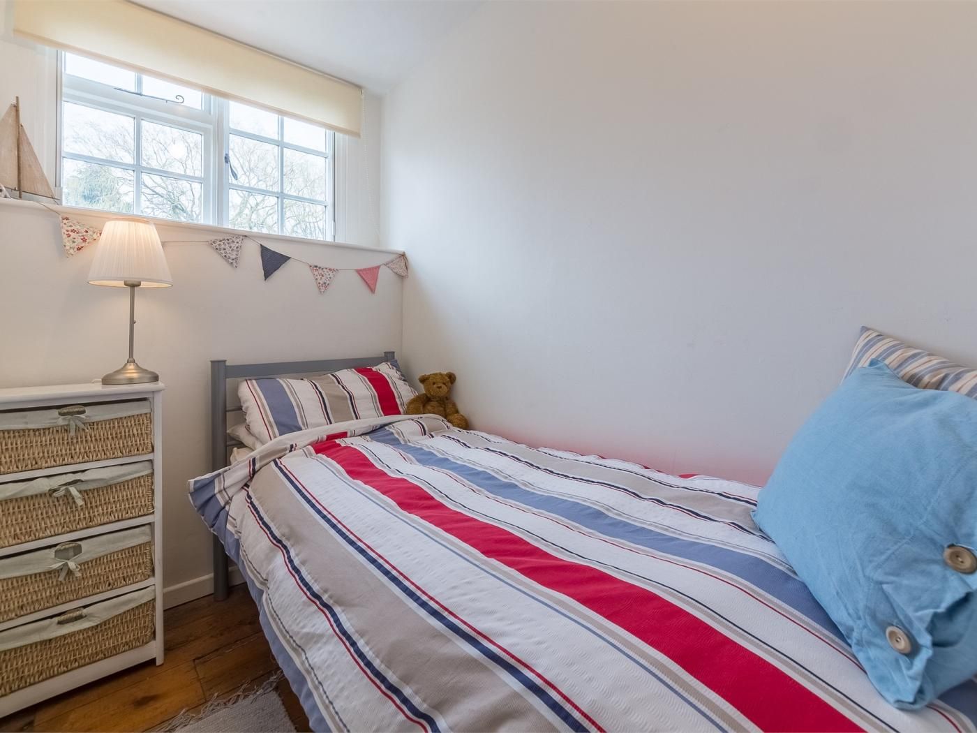 A bedroom with a bed, a bedside lamp, a teddy bear, a dresser with baskets, and a window with a view.