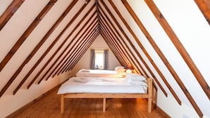 Une pièce avec un plafond en pente, des poutres en bois, un lit avec du linge de lit blanc, une fenêtre avec des rideaux et une prise électrique au mur.