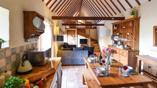 Une cuisine rustique avec des poutres en bois, une table à manger dressée avec des assiettes et des verres, et un plan de travail avec une bouilloire et des légumes.