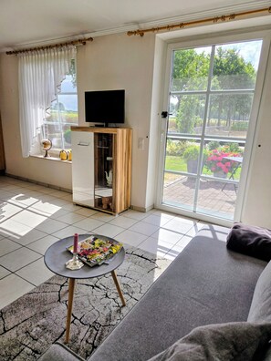 Ein Wohnzimmer mit einer Couch, einem kleinen Tisch mit Kerze, einem Fernseher und einem Fenster mit Vorhängen.