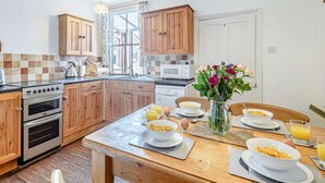 A kitchen with wooden cabinets, a stove, oven, and a dining table set with breakfast. - 3 Bed in Thornham (oc-kt022) (Thornham)
