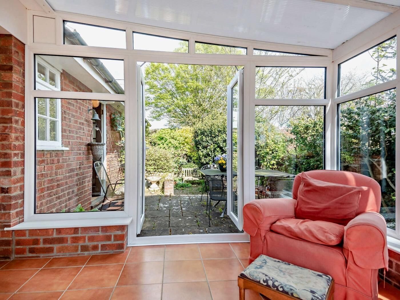 A conservatory with a red brick wall, a pink chair, and a view of a garden.