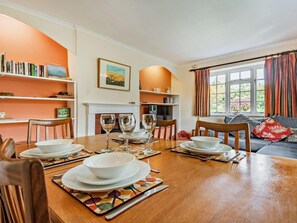 Een eetruimte met een houten tafel gedekt voor vier personen, omringd door stoelen. Achtergrond met boekenkasten en een open haard.