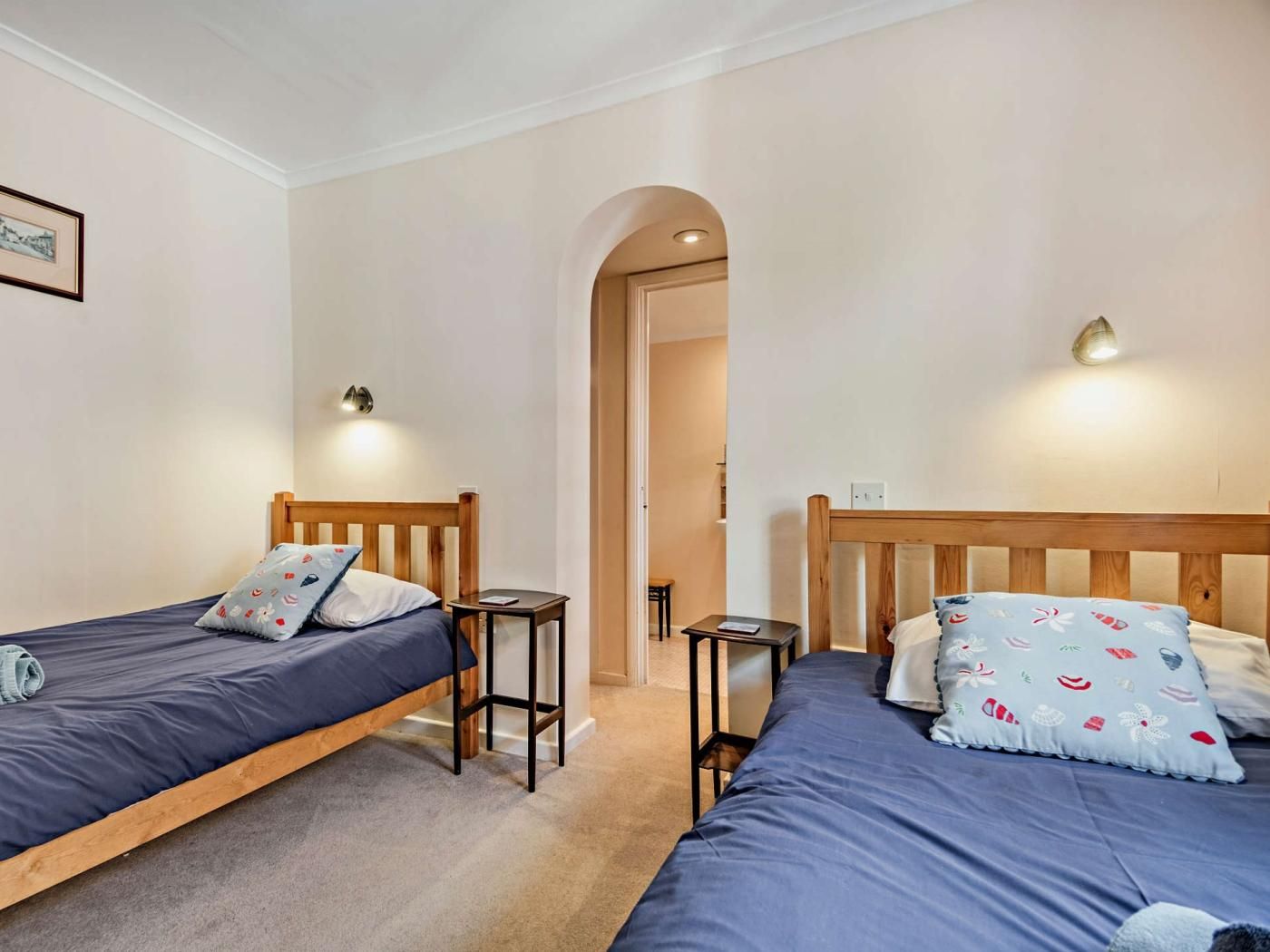 A room with two single beds, each with a wooden headboard and a blue mattress. There are two bedside tables, one on each bed, and a small table between the beds. The room has a neutral color scheme with beige walls and a carpeted floor.
