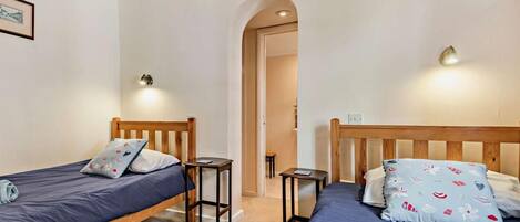 A room with two single beds, each with a wooden headboard and a blue mattress. There are two bedside tables, one on each bed, and a small table between the beds. The room has a neutral color scheme with beige walls and a carpeted floor.