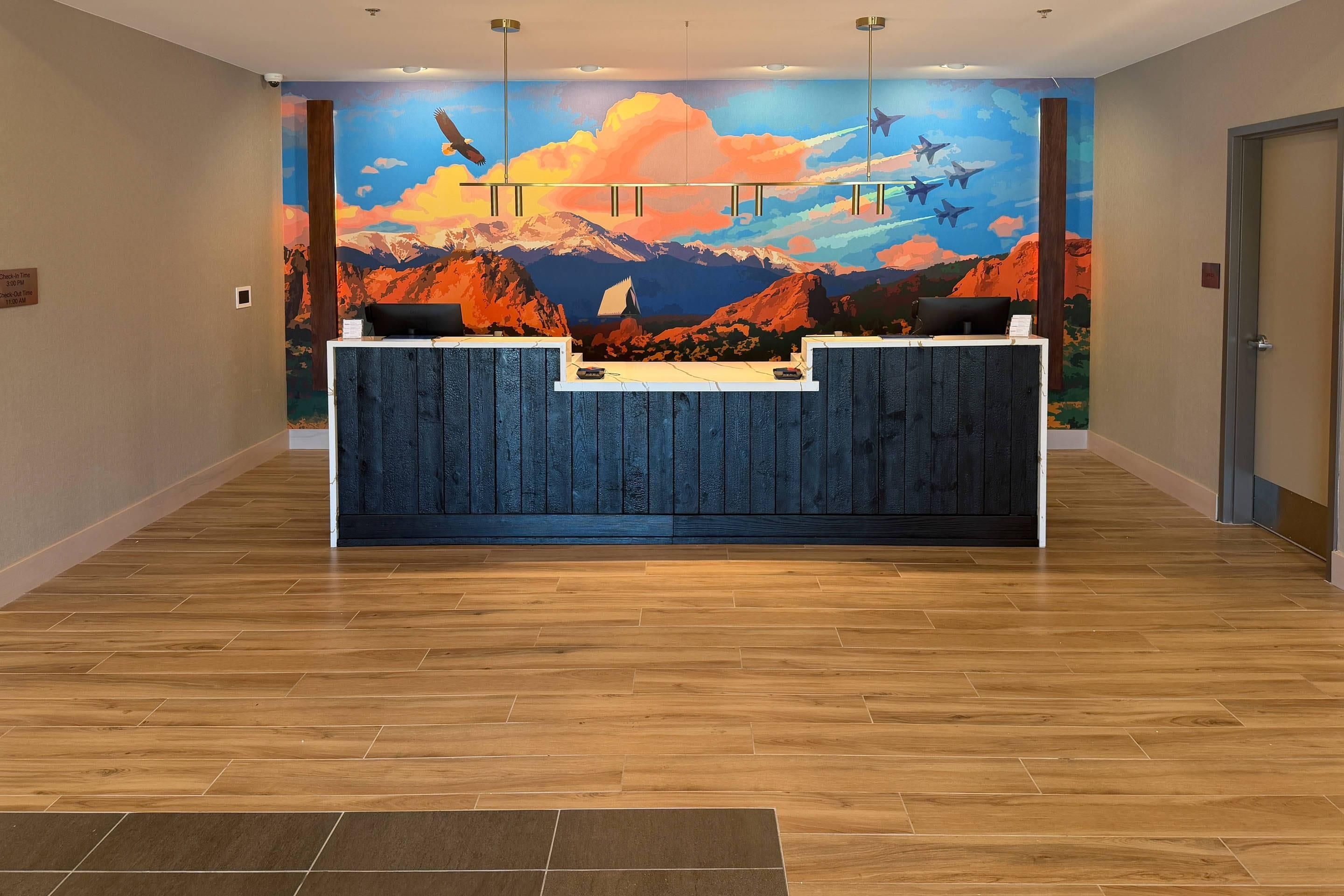 Un mostrador de recepción con un mural de montañas y aviones, suelo de madera y un cartel de registro de entrada/salida.