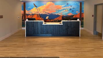Un mostrador de recepción con un mural de montañas y aviones, suelo de madera y un cartel de registro de entrada/salida.