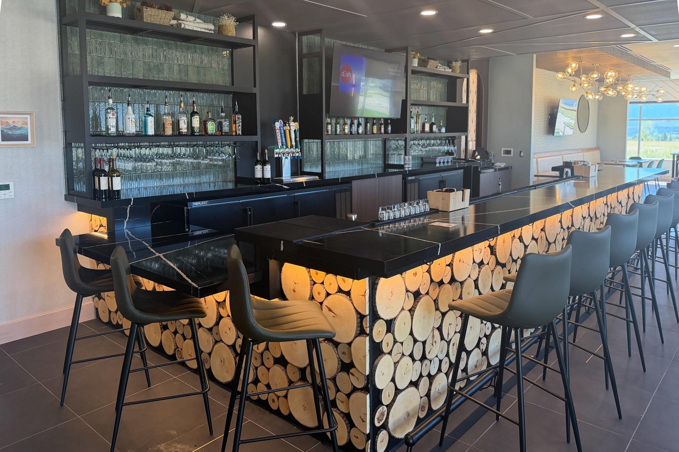 Zona de bar con una barra larga, varias opciones de asientos y una variedad de botellas y vasos en estanterías.