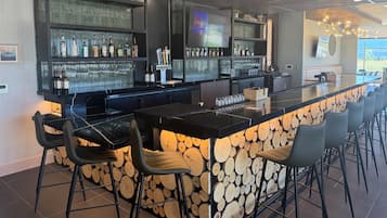Zona de bar con una barra larga, varias opciones de asientos y una variedad de botellas y vasos en estanterías.