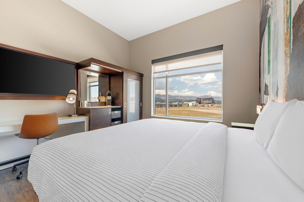 A modern hotel room with a large bed, a desk, a chair, and a window offering a view of the cityscape.
