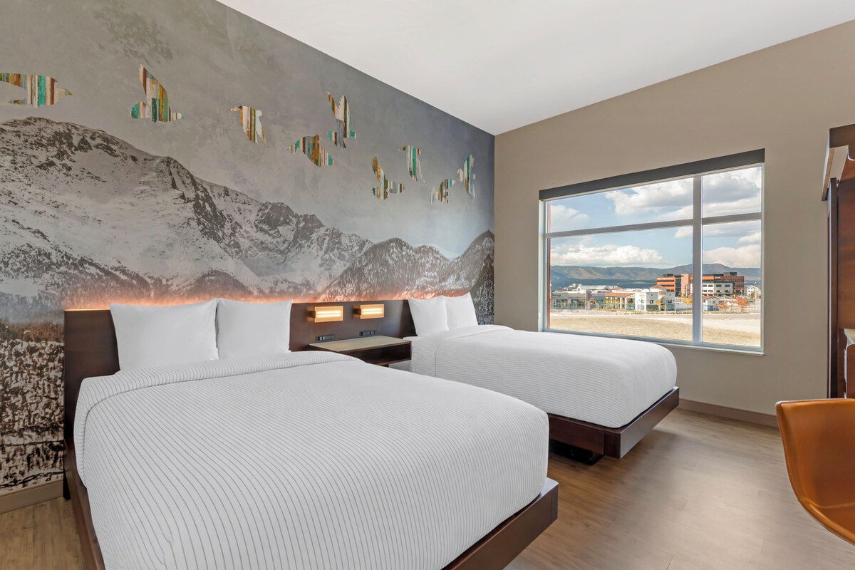 A modern hotel room with two beds, a large window with a city view, and a mountain-themed wall mural.
