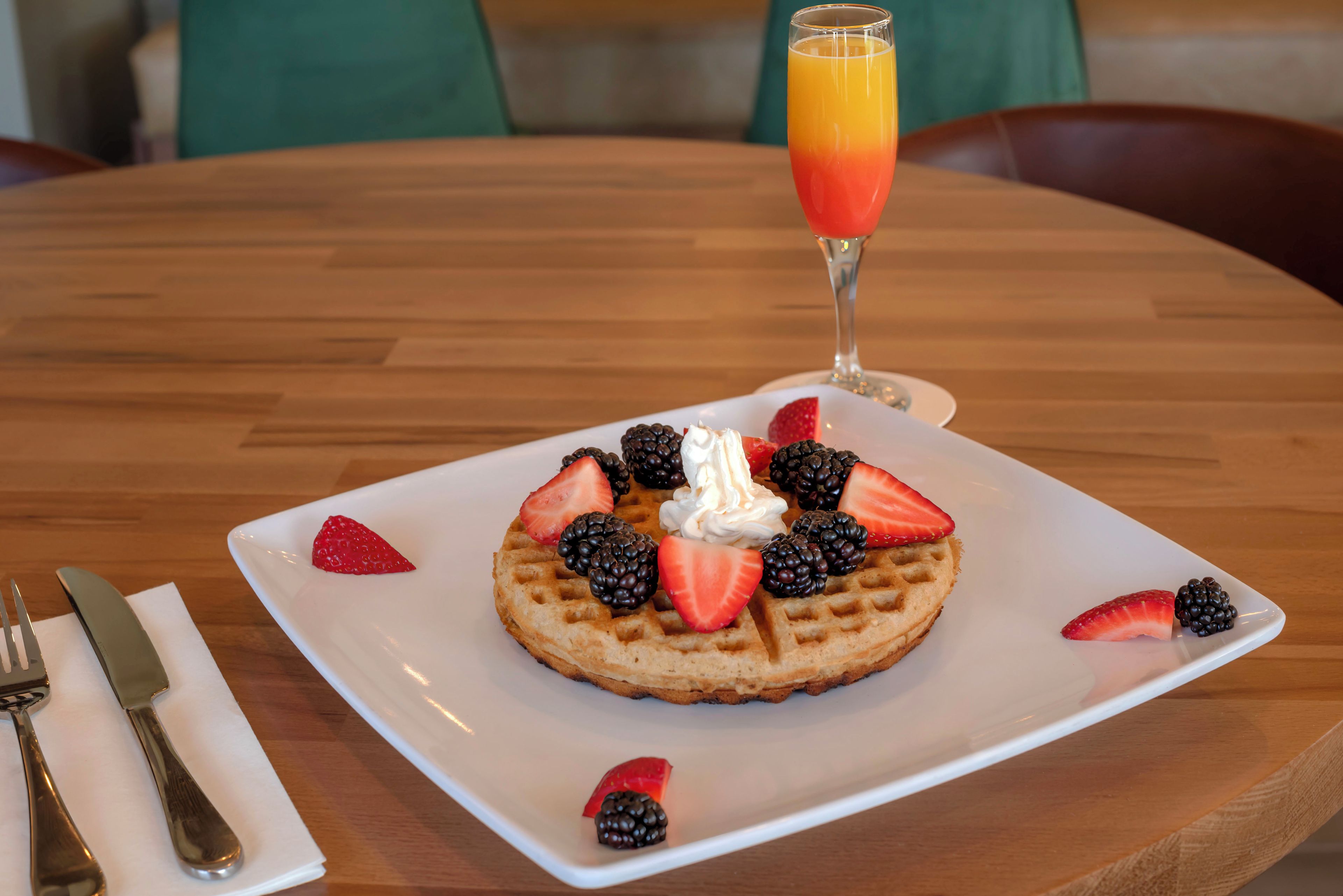 Ein Teller mit Waffeln, Schlagsahne und Beeren, ein Glas Orangensaft und Besteck auf einem Holztisch.