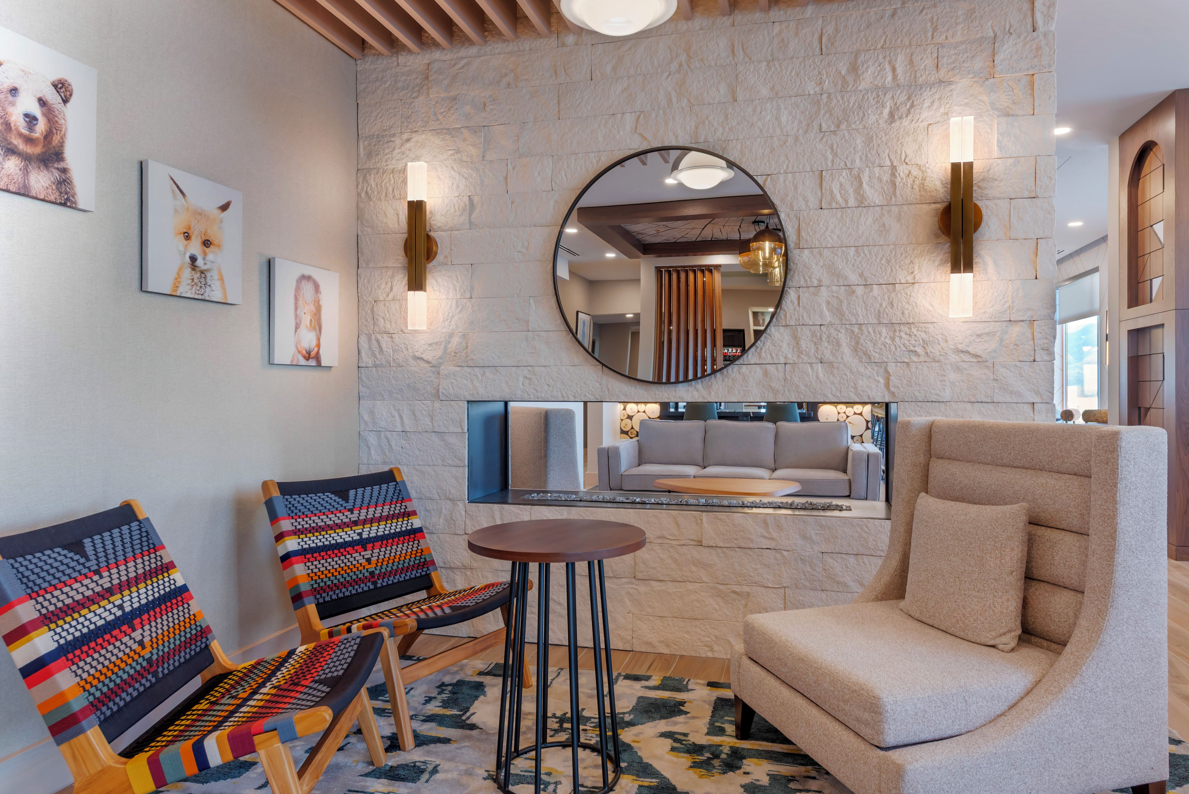 A modern lounge area with a stone wall, two colorful woven chairs, a round table, and a large armchair.