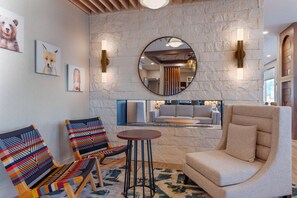 A modern lounge area with a stone wall, two colorful woven chairs, a round table, and a large armchair. - Cambria Hotel Colorado Springs - Air Force Academy (Colorado Springs)