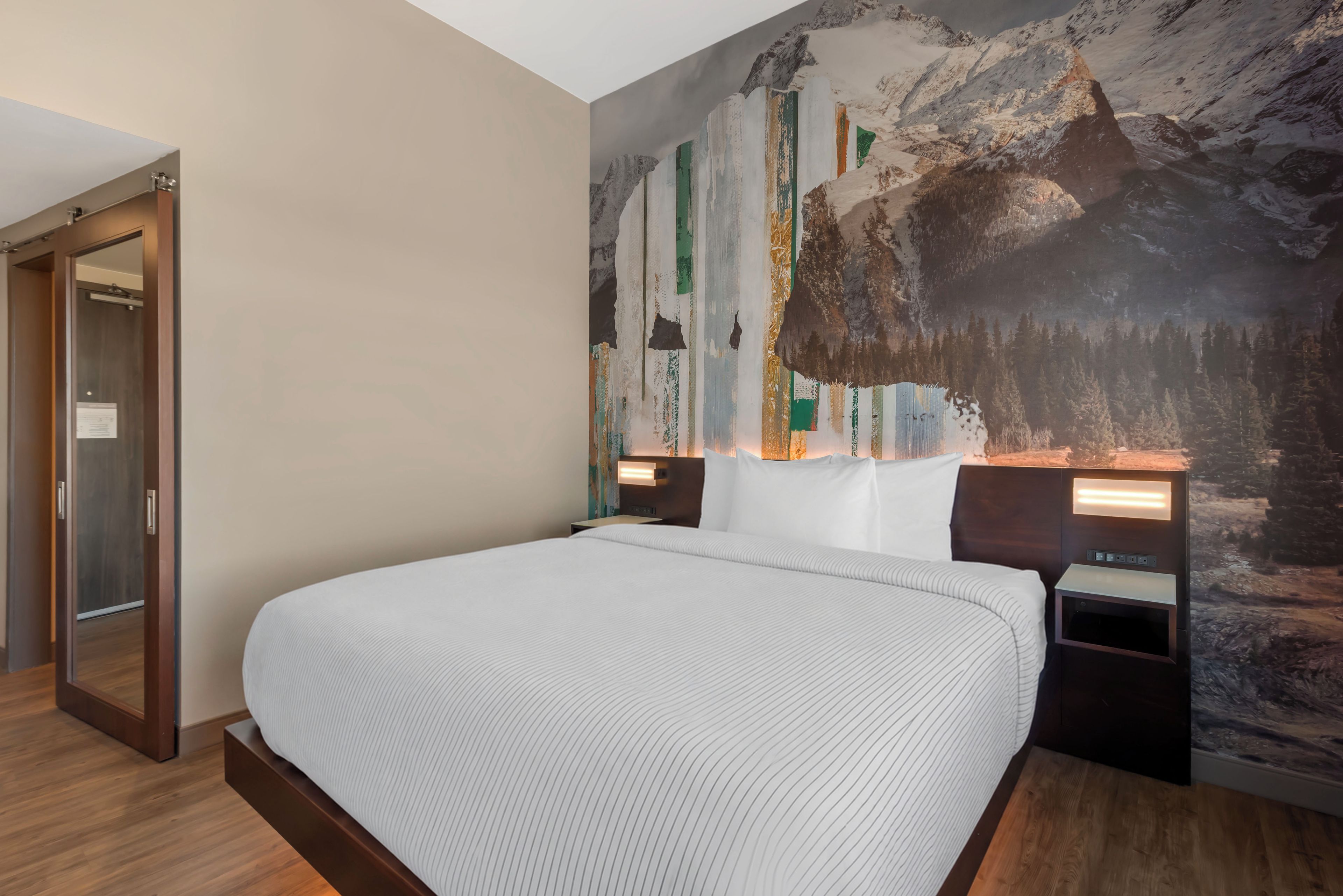 Une chambre d’hôtel avec un grand lit, un plancher en bois, une fresque murale sur le thème de la montagne et une porte dotée d’un panneau en verre.