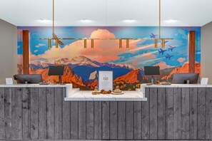 A reception desk with two computer monitors, a wooden reception desk, and a mural of mountains and airplanes. - Cambria Hotel Colorado Springs - Air Force Academy (Colorado Springs)