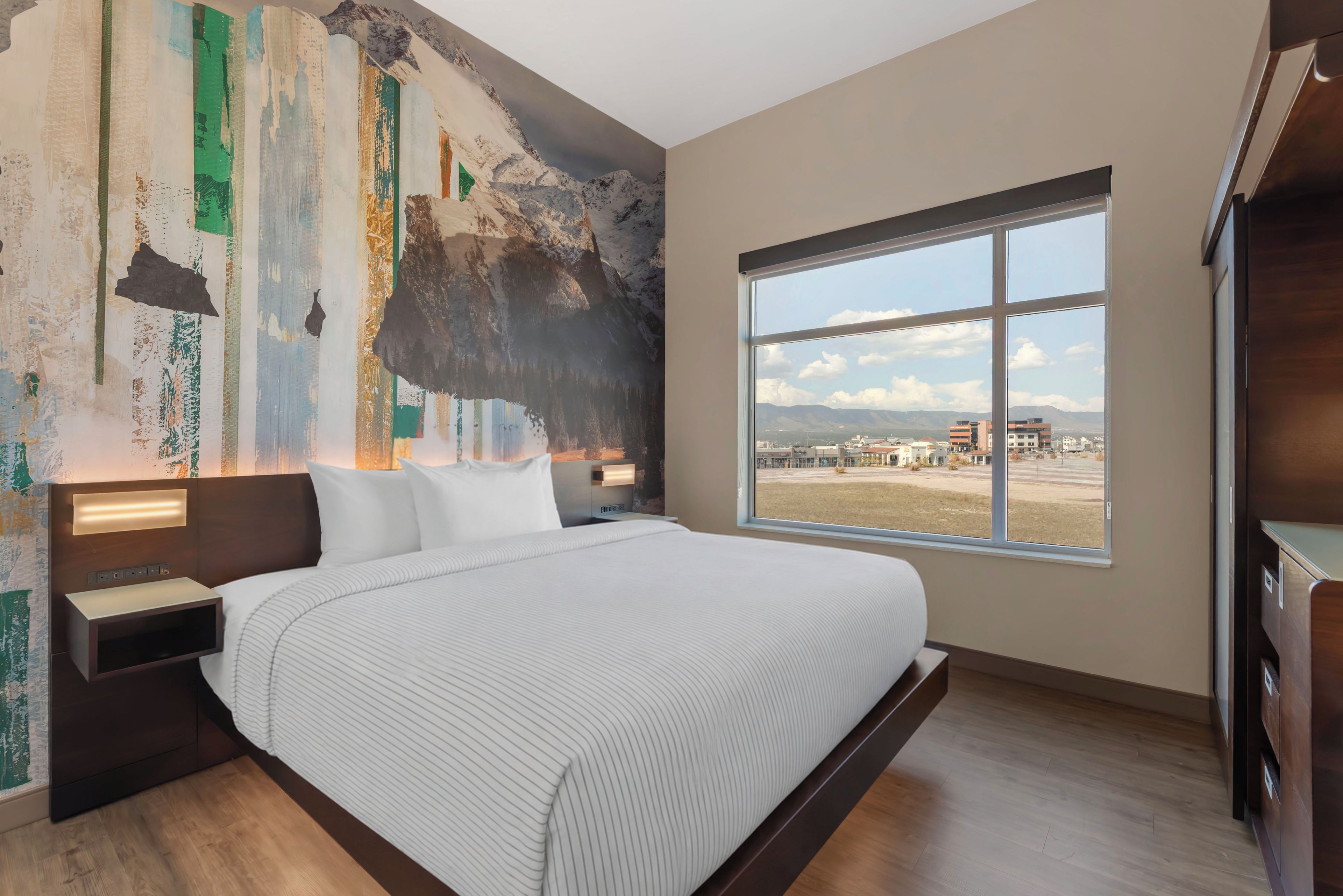 Ein Hotelzimmer mit einem großen Bett, Blick auf die Stadt durch ein großes Fenster und einer Wand mit Wandgemälde.