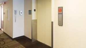 Pasillo de hotel con piso alfombrado, un teléfono, un detector de incendios y un panel de control de ascensor.