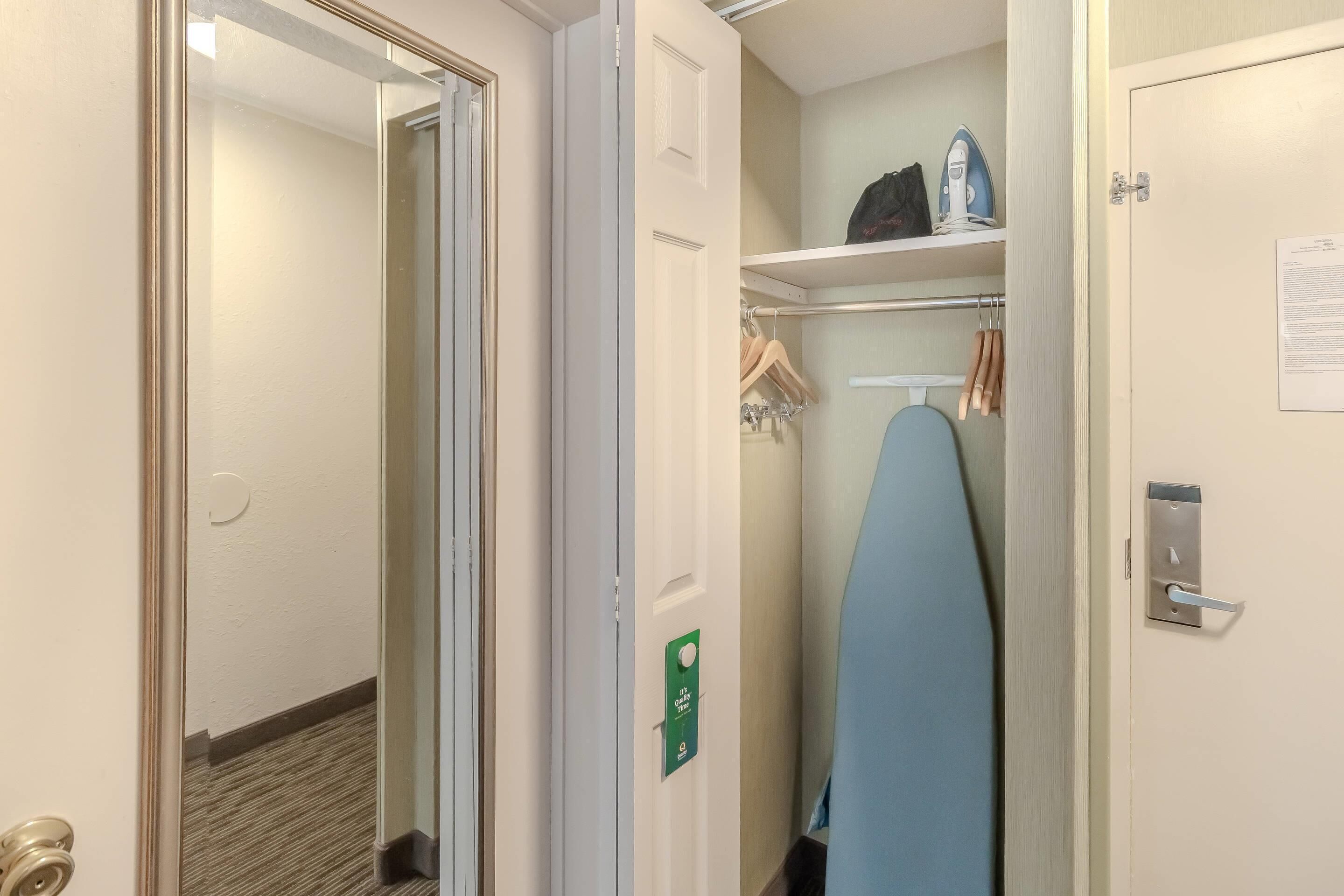 Habitación de hotel con una puerta que da a un pasillo, un armario con ropa colgada y una tabla de planchar azul.