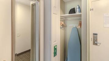 Habitación de hotel con una puerta que da a un pasillo, un armario con ropa colgada y una tabla de planchar azul.