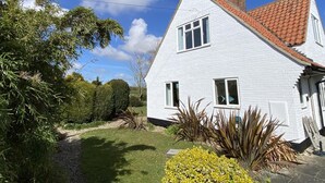Ein weißes, zweistöckiges Haus mit rotem Ziegeldach, einem sichtbaren Schornstein und einem kleinen Garten mit verschiedenen Pflanzen und einem Kiesweg.