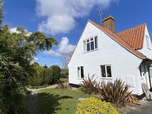 Ein weißes, zweistöckiges Haus mit rotem Ziegeldach, einem sichtbaren Schornstein und einem kleinen Garten mit verschiedenen Pflanzen und einem Kiesweg.