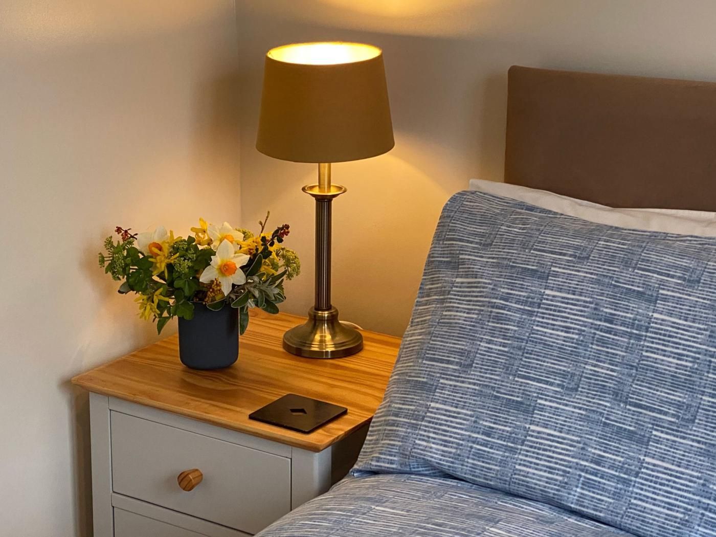 A neatly made bed with a blue patterned pillow, a bedside table with a lamp and a flower arrangement, and a drawer with a wooden knob.