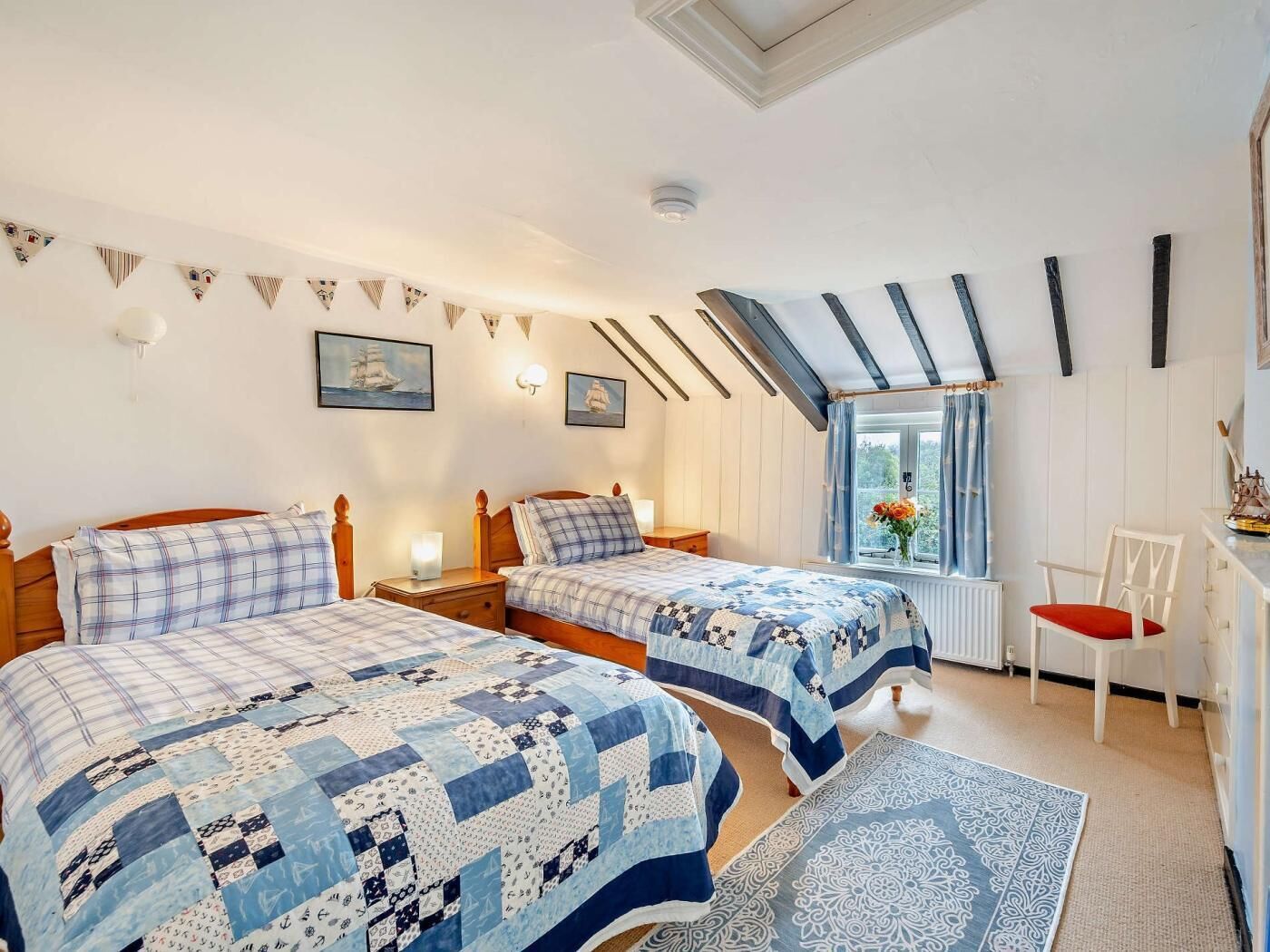A bedroom with two beds, a skylight, a window with curtains, a chair, and a small table.