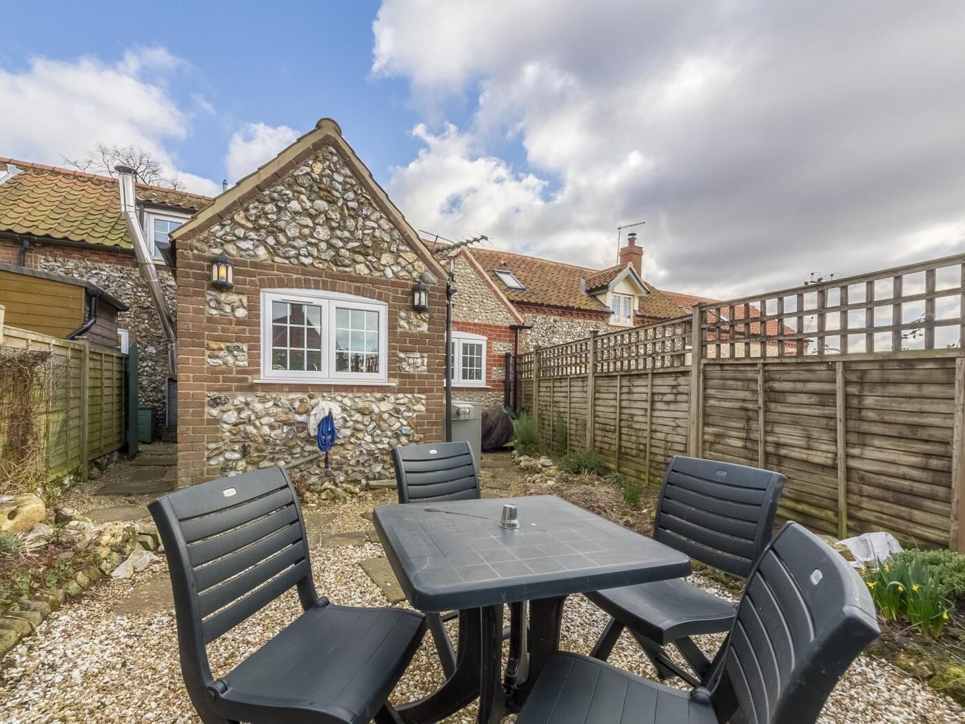 A stone house with a tiled roof, a wooden fence, and a gravel patio with four black chairs and a table.