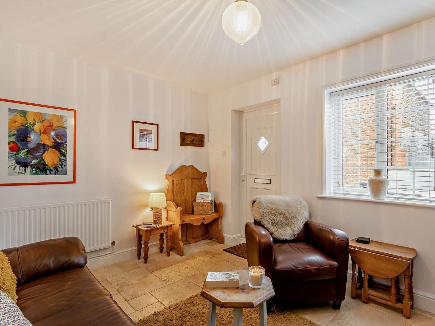 Un salon chaleureux avec un canapé en cuir marron, une chaise en bois, une petite table avec une bougie et une fenêtre avec des stores.