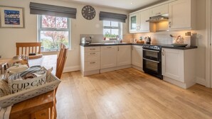 A kitchen with white cabinets, a black stove, and a wooden dining table with a basket labeled 'E-CUP CAKE'. - 3 Bed in Chathill (oc-62640) (Swinhoe)