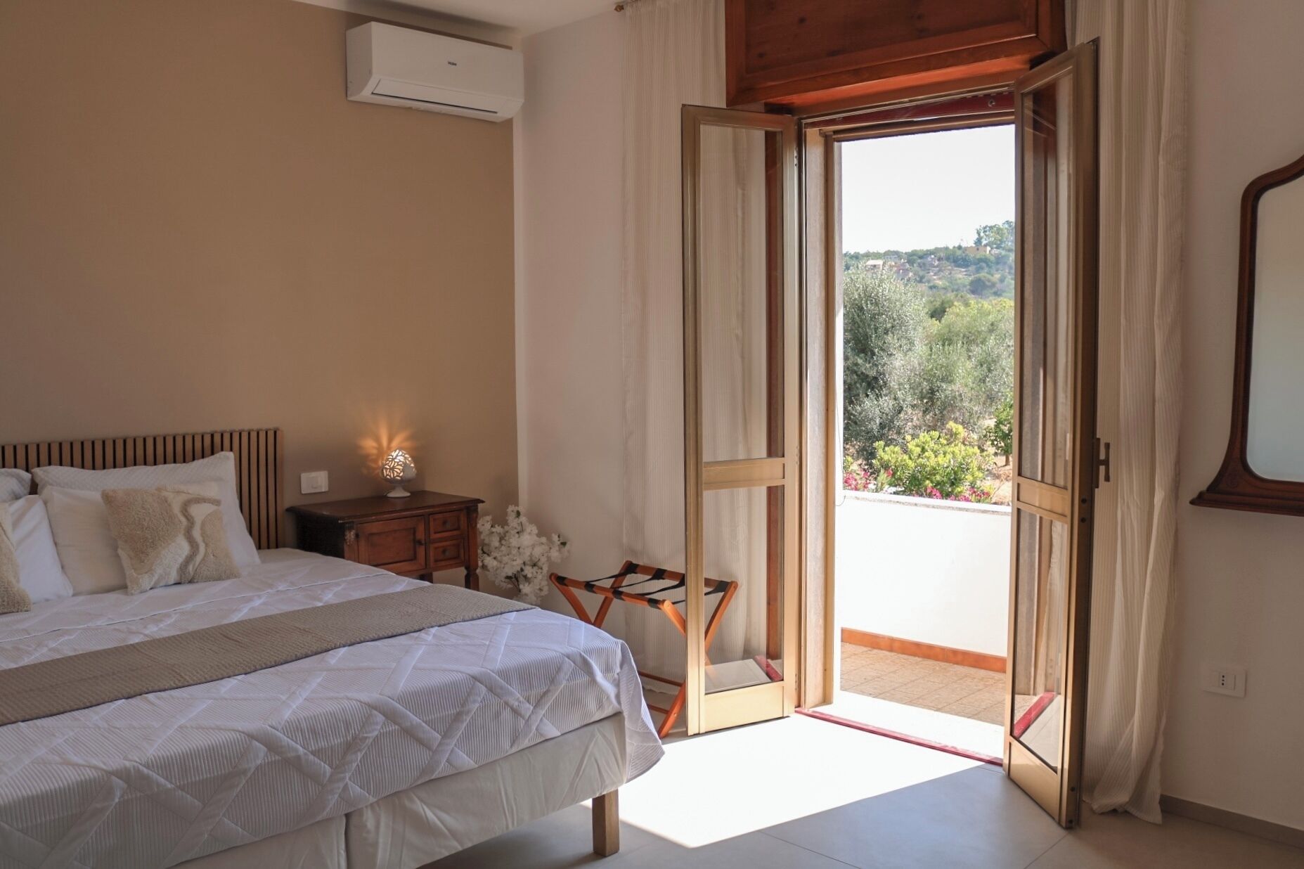Una camera da letto con un letto, un comodino, una lampada e una porta aperta che dà su un balcone con vista sul verde.